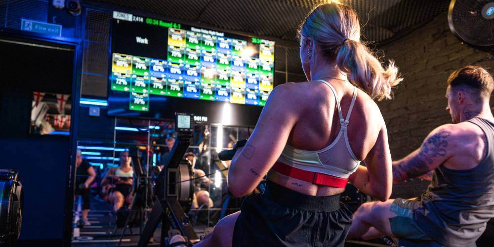 A woman and a man on rowing machines during a workout, in front of a screen full of Myzone heart rate tiles