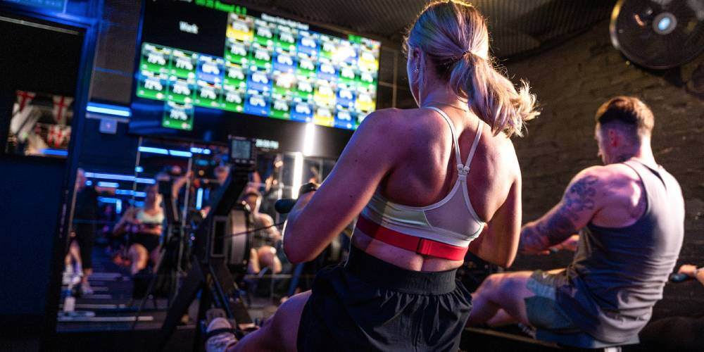 Two gym members on rowing machines in front of a bright Myzone heart rate tile screen