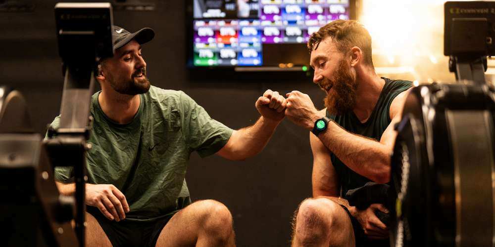 Two people in the gym, fist bumping in front of a Myzone screen